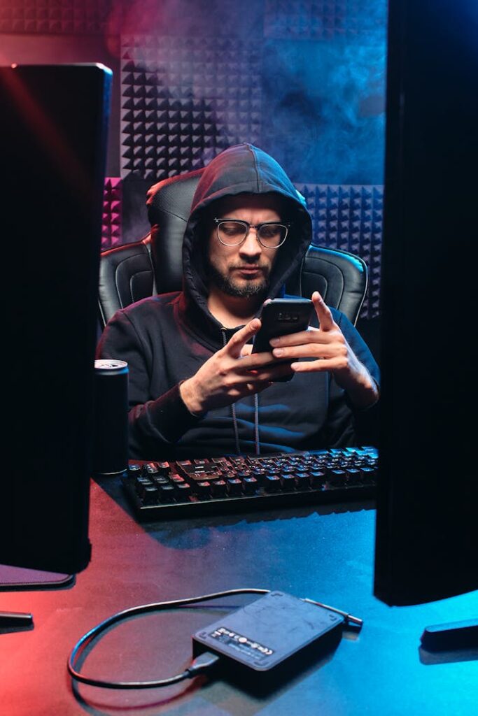 A man in a hoodie uses a smartphone at a computer desk, highlighting a tech atmosphere.