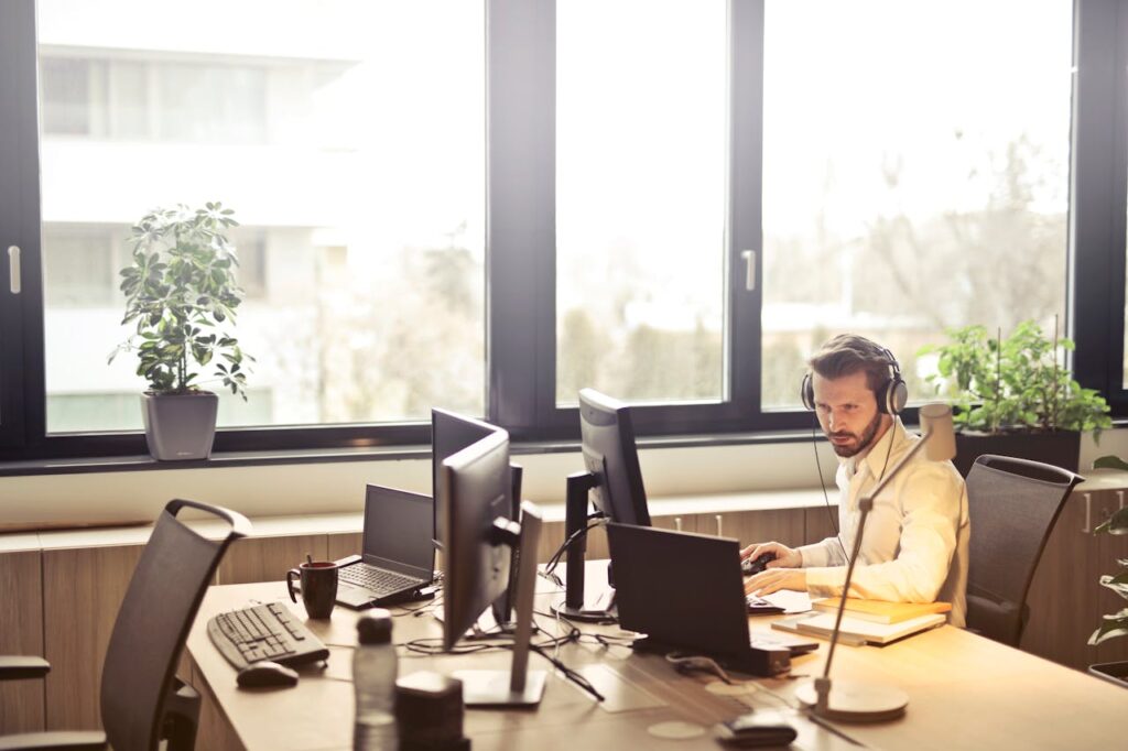 pexels-photo-845451 A businessman sits at a desk using multiple computers and a headset in a well-lit modern office.